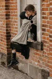 tragetuch aus leinen - Frau sitzt in Fensterbank mit Baby im Tragetuch aus Leinenstoff