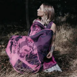 A woman sits relaxed in the forest and wears a purple-pink baby sling made of 100% cotton with a large elephant motif.