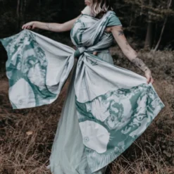 Woman carries her baby in the autumnal reed grass on her back. The long, light green baby sling with an elephant motif falls down elegantly like a dress.