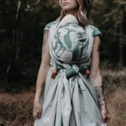 Une femme porte son bébé de manière ergonomique devant le ventre dans une écharpe vert clair avec un grand visage d'éléphant tissé.